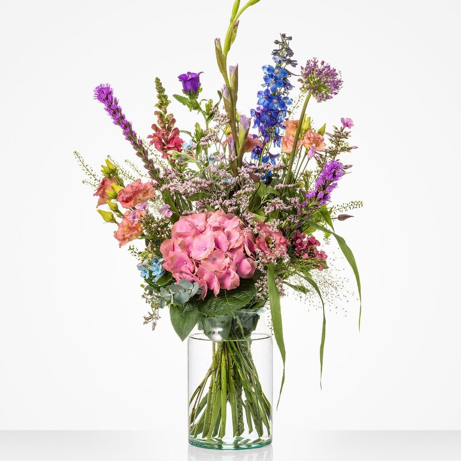 Middelgroot gekleurd boeket in een glazen vaas met diverse bloemen, waaronder roze hortensia’s, blauwe en paarse bloemen.