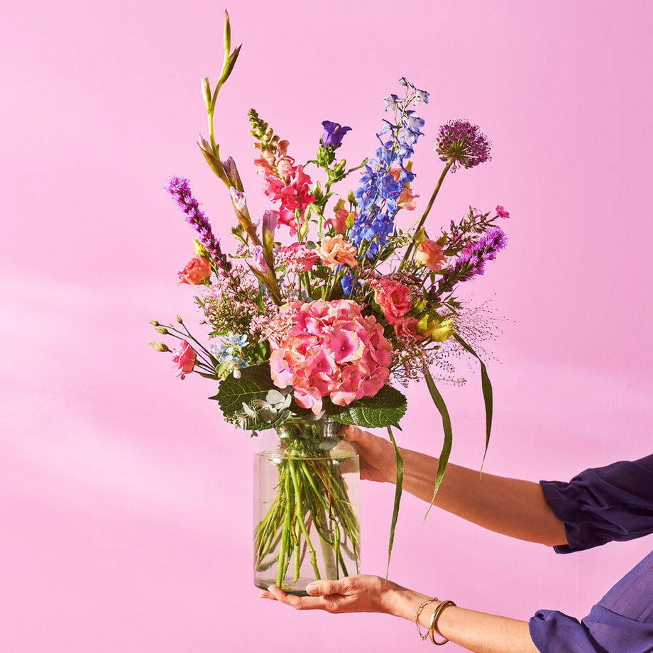 Groot gekleurd boeket in een glazen vaas op een houten kast tegen een roze muur, vastgehouden door een persoon in een blauwe blouse.