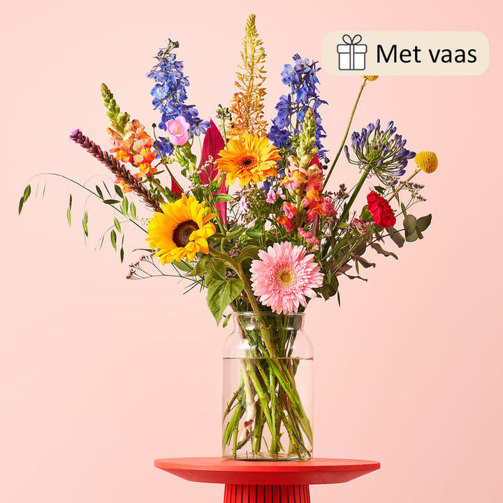 Sfeerfoto van een boeket in een glazen vaas op een rode tafel met een lichte achtergrond. Het boeket bevat zonnebloemen, gerbera’s, leeuwenbekjes, en andere kleurrijke bloemen voor een zomerse uitstraling.