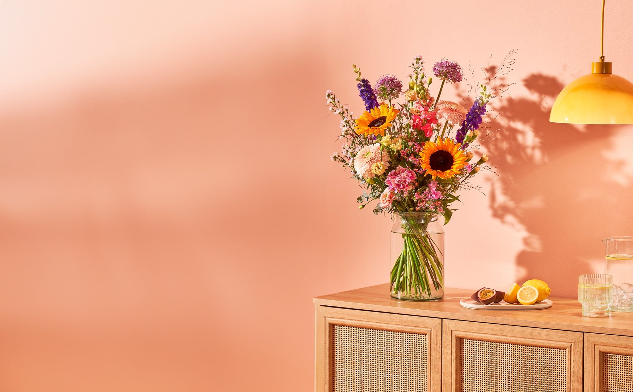 Vrolijk boeket met kleurrijke bloemen, bezorgd door Buurtbloemist, gestyled in een glazen vaas op een houten kast.